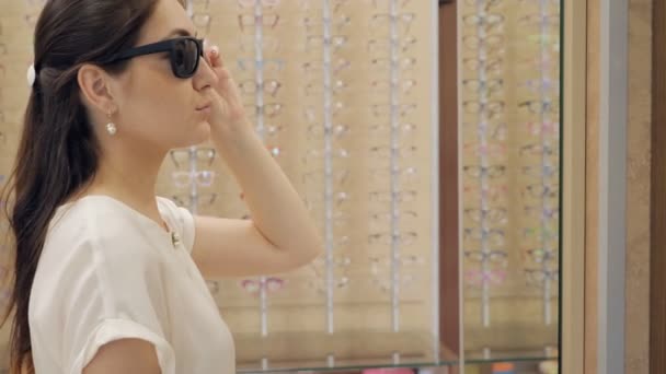 Mulher em uma loja de óptica experimentando óculos de sol na frente de um espelho — Vídeo de Stock