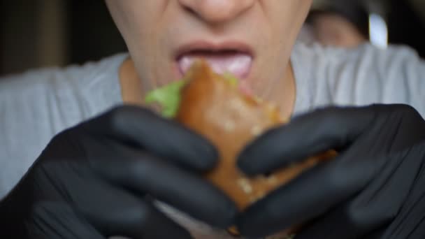 Крупным планом неизвестного человека в черных перчатках, поедающего бургер в кафе — стоковое видео