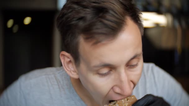 Jovem de homem com luvas pretas comendo um hambúrguer em um café . — Vídeo de Stock