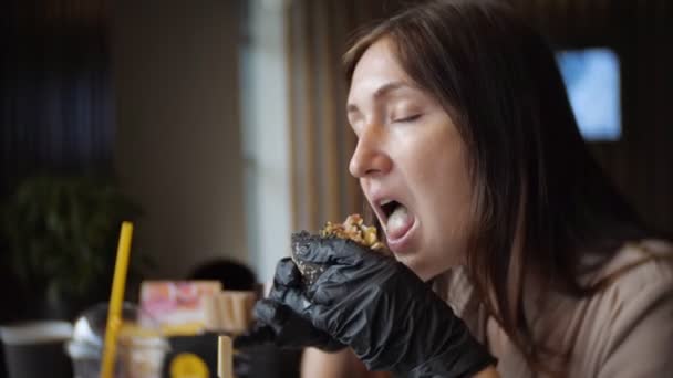 Jeune fille affamée en gants noirs mangeant un hamburger juteux dans un café — Video