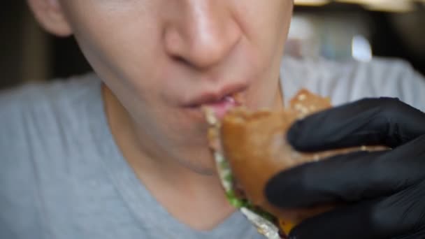 Hungrig man i svarta handskar äta en hamburgare på ett café — Stockvideo