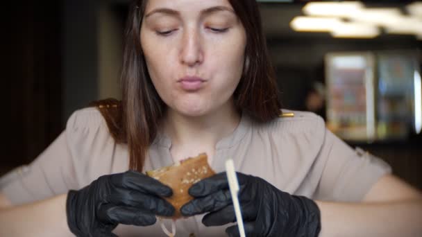 饥饿的女孩在黑手套吃一个多汁的汉堡在咖啡馆里 — 图库视频影像