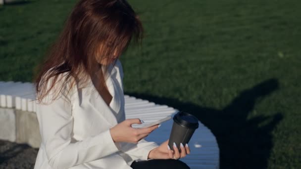 Ung kvinna med smartphone som sitter i parken. Fashion business foto av vacker flicka i vit casual suite med telefonen och kopp kaffe — Stockvideo
