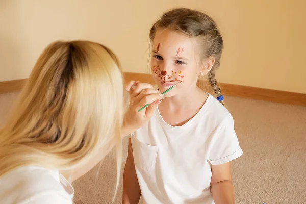 Возбужденная маленькая девочка с раскрашенной мордой развлекается с мамой, рисующей на лице кисточкой — стоковое фото
