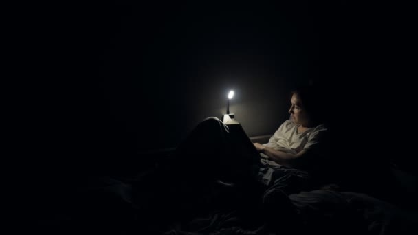 Молодая девушка читает книгу в постели ночью с лампой света — стоковое видео