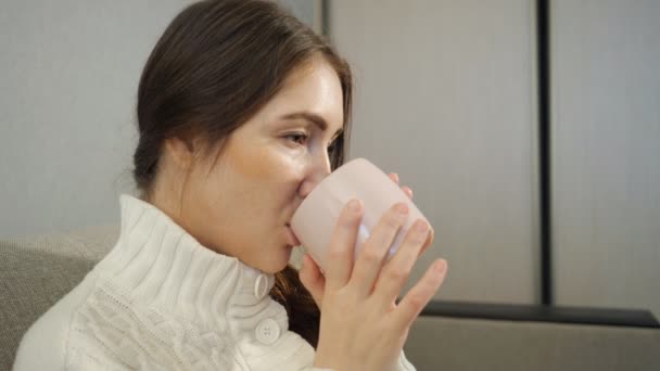 Mladá žena v bílém svetru sedí na pohovce a a pití kávy doma — Stock video