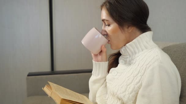 Mooi meisje het lezen van een boek en drinken koffie op de Bank thuis — Stockvideo