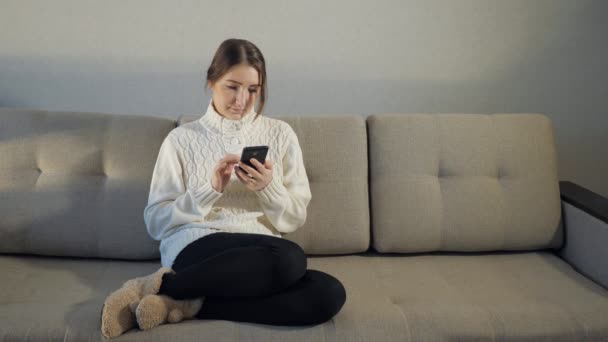 Όμορφο κορίτσι στο πουλόβερ που κάθεται στον καναπέ και να διαβάσετε τα μηνύματα στο τηλέφωνο — Αρχείο Βίντεο