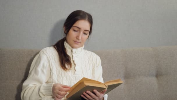 Όμορφη νεαρή γυναίκα που κάθεται στον καναπέ και την ανάγνωση του βιβλίου στο σπίτι — Αρχείο Βίντεο