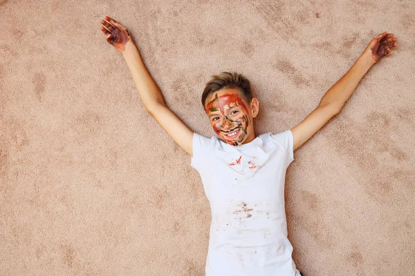 Niño pequeño con palmas y la cara en pinturas de colores brillantes mirando a la cámara tumbada en el suelo — Foto de Stock