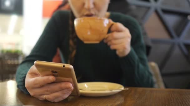 Неузнаваемая женщина в зеленом свитере с телефоном в руке, пьющей напиток . — стоковое видео