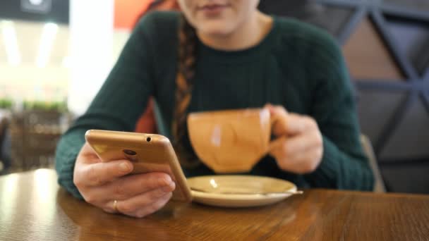 Телефон в руке девушки в фокусе, женщина пьет кофе на размытом фоне — стоковое видео