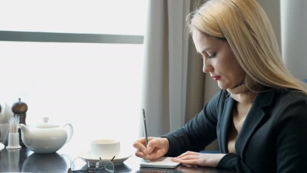 Giovane ragazza che fa i compiti, scrivere appunti in blocco note seduto nel caffè — Video Stock