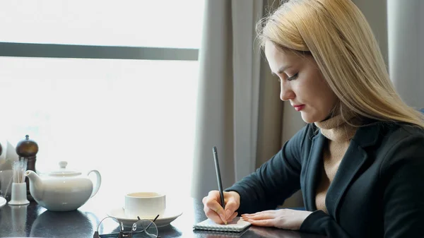 Молодая девушка делает домашнее задание, пишет заметки в блокноте сидя в кафе — стоковое фото
