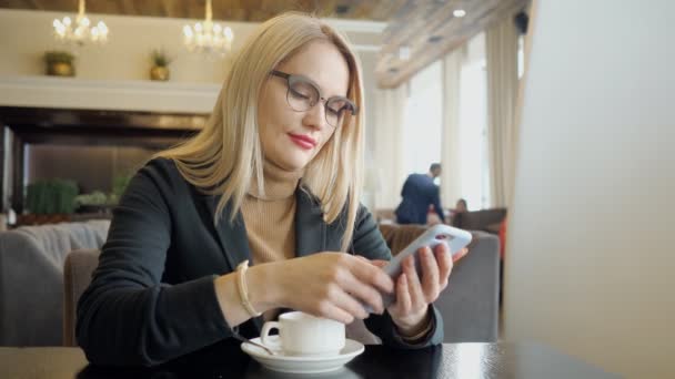 Chica rubia en traje con teléfono sentado en una mesa en un café — Vídeos de Stock