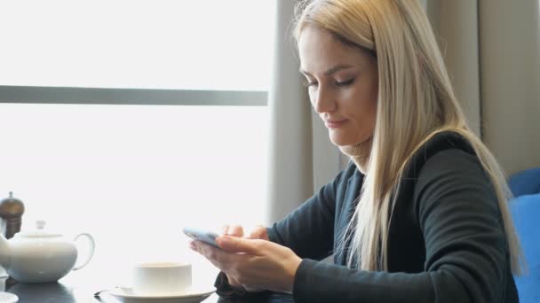 Krásná mladá blondýnka s telefonem sedí u stolu v kavárně — Stock video