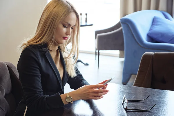 Mujer de negocios rubia en traje con teléfono sentado en una mesa en un café —  Fotos de Stock