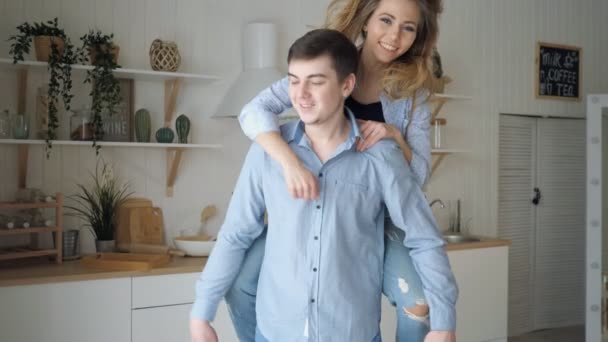 Meisje sprong op de schouders van de jongen lachen kussen man in de keuken — Stockvideo