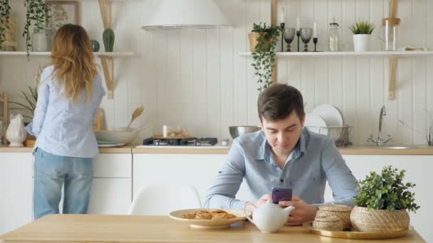 Ragazza mette biscotti tazze sul ragazzo tavolo guarda nel telefono — Video Stock
