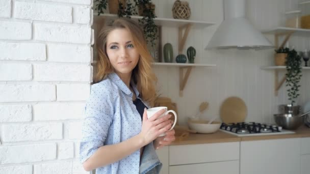 Menina loira bebe café e sorri perto da janela na cozinha — Vídeo de Stock