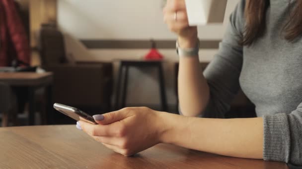 Femme boit une tasse de café dans le café et de lire quelque chose dans le téléphone mobile. Mains avec téléphone gros plan . — Video