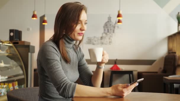 Chica joven en la cafetería está escribiendo un mensaje en el teléfono y beber una taza de café — Vídeos de Stock