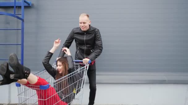 Guy rolls his attractive young girlfriend in the trolley on parking lot of the supermarket. — Stock Video