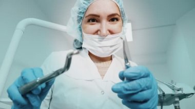 Diş hekimi kadın doktor elinde diş aletleri forseps ve iğne tutuyor ve kamera bakıyor.