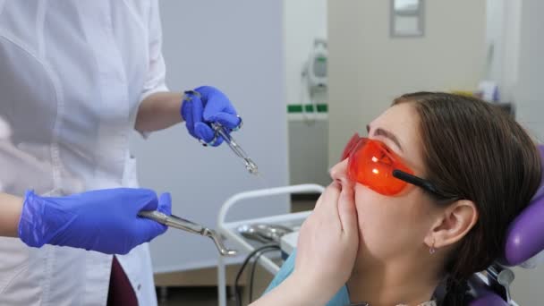 Молодая женщина боится удалить зуб сидя в стоматологическом кресле, в то время как врач стоит рядом с пациентом, держа щипцы в руках . — стоковое видео