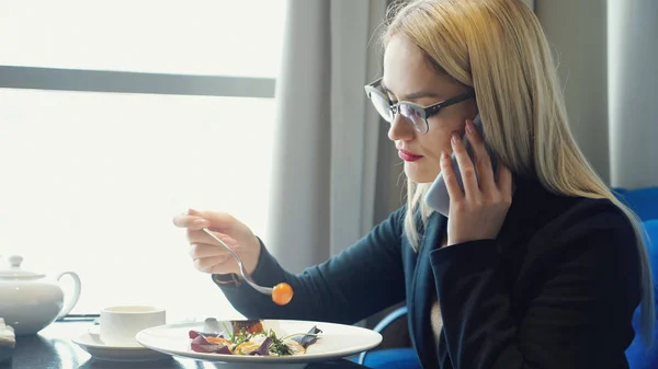 Молода жінка менеджер розмовляє по телефону і їсть перерву під час перерви в кафе — стокове фото