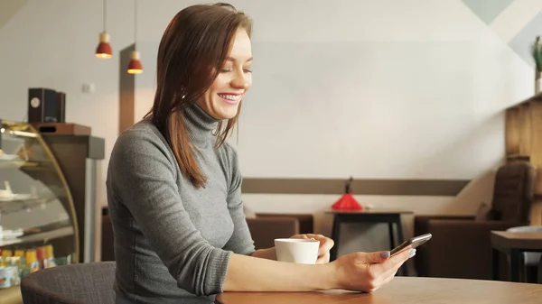 Junges Mädchen im Café tippt eine Nachricht am Telefon und trinkt eine Tasse Kaffee — Stockfoto