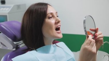Dişçi koltuğundaki genç kadın, tedaviden sonra dişlerini aynada inceliyor. Diş bakımı konsepti.