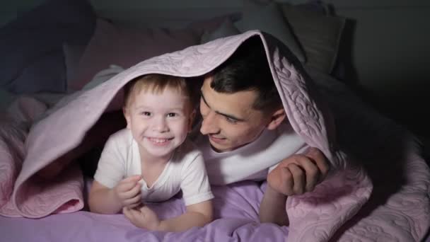 Papá y su pequeño hijo están jugando a esconderse bajo la manta en la oscuridad . — Vídeo de stock