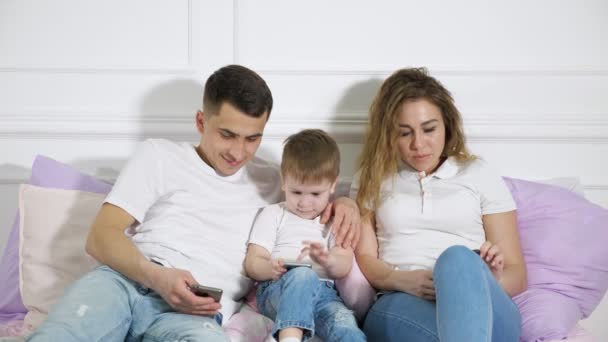 Rodzina z dzieckiem szukają w swoich telefonach komórkowych kładąc na łóżku. Uzależnienie od gadżety. — Wideo stockowe