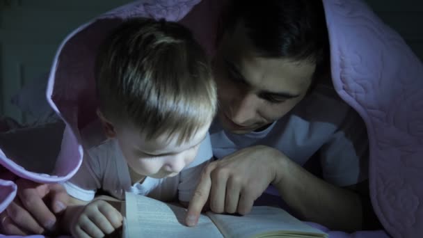 Папа вчить свого маленького сина, щоб прочитати книгу ховається під ковдру. — стокове відео