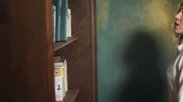 Γραφείο γυναίκα επιλέγει ένα βιβλίο για την ανάγνωση στέκεται κοντά στη βιβλιοθήκη. — Αρχείο Βίντεο