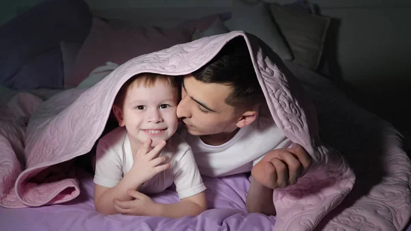 Dad and his little son is playing hiding under the blanket in dark.