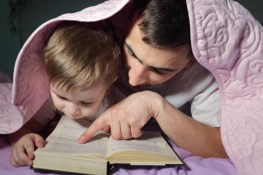Babam küçük oğluna battaniyenin altında saklanan bir kitap okumayı öğretir..