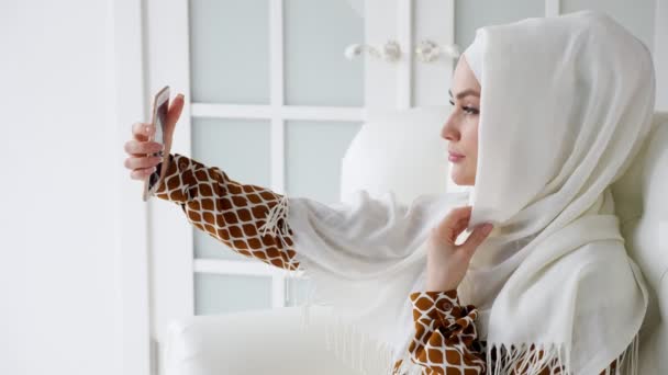 Young muslim woman in hijab is making selfie on smartphone sitting on the sofa. — 비디오