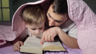 Babam küçük oğluna battaniyenin altında saklanan bir kitap okumayı öğretir..