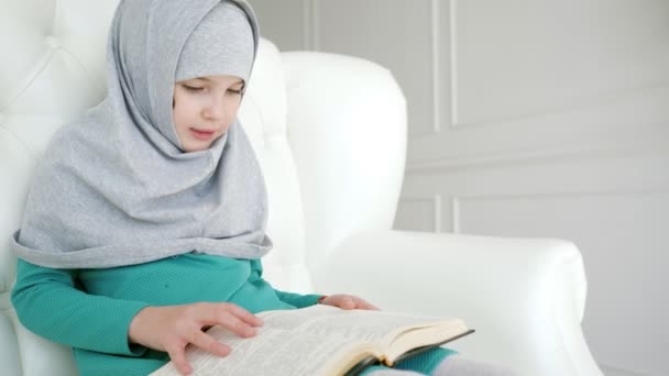 Мусульманська дівчина дитина в хіджабі читає книгу сидячи на дивані будинку. — стокове відео