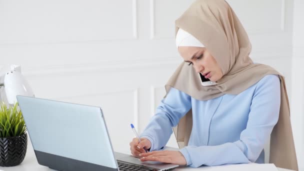 Muslim kvinna snackar med klienten på rörlig telefonerna, tillverkningen papper anteckningen och sett på laptop skärm. — Stockvideo