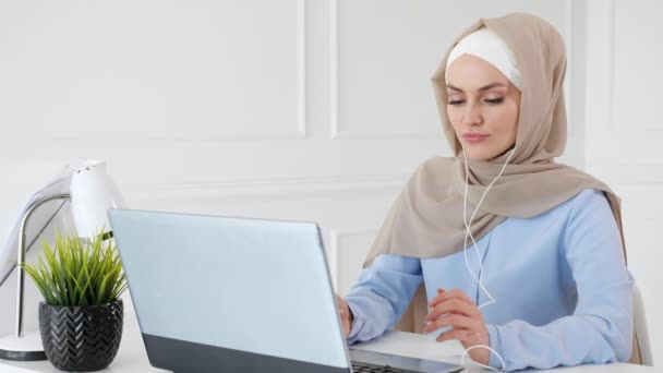 Mujer musulmana joven está escuchando música en los auriculares usando el ordenador portátil y disfrutando de ella . — Vídeos de Stock