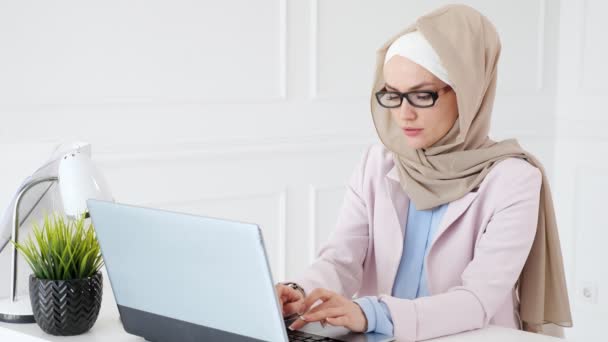 Mujer en hijab y gafas está haciendo un trabajo aburrido en su computadora portátil . — Vídeo de stock