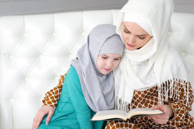 Muslim mom in hijab is reading her little daughter a book sitting on the sofa. clipart