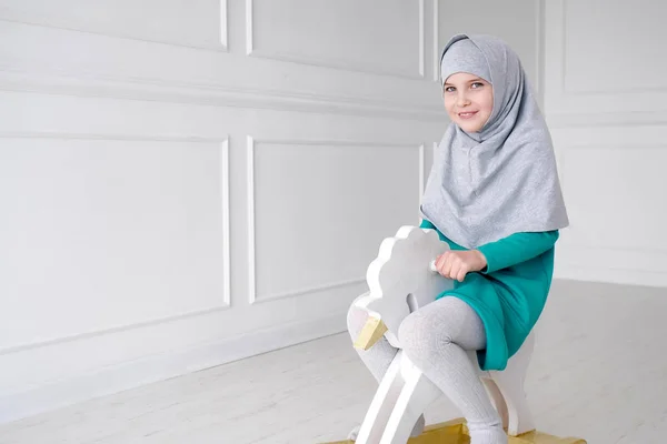 穆斯林少女在头巾和礼服是玩骑在玩具马摇椅在她的房间. — 图库照片