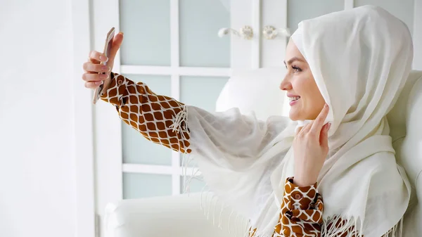 穿着头巾的年轻的穆斯林妇女正坐在沙发上用智能手机自作主张. — 图库照片