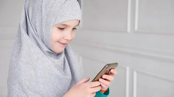 灰色のヒジャーブで若いイスラム教徒の十代の少女の肖像画は、スマートフォンを再生しています — ストック写真