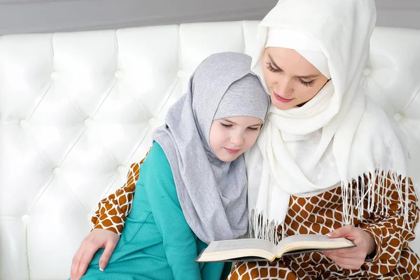 Muslim mom in hijab is reading her little daughter a book sitting on the sofa.