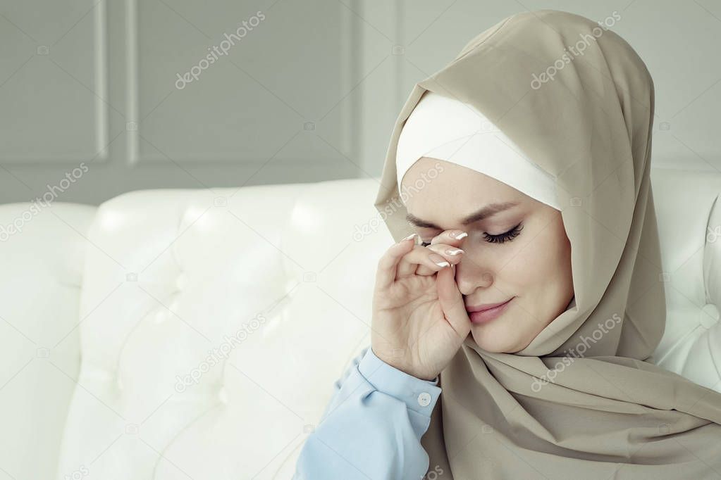 Sad crying muslim woman in hijab is sitting on the sofa at home.